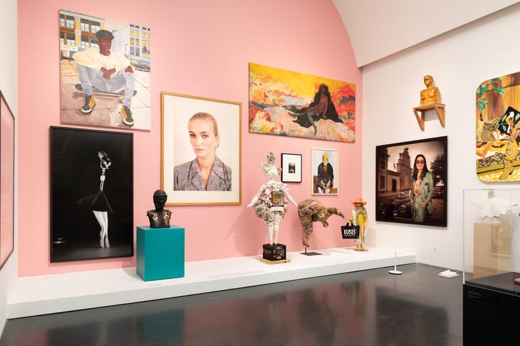 An installation image of the show. There are two walls, one is pink and the other is white, which show various paintings, photographs, sculptures, and drawings included in the show. In the center of the image, there is a photograph of a white feminine person looking directly at the camera. 