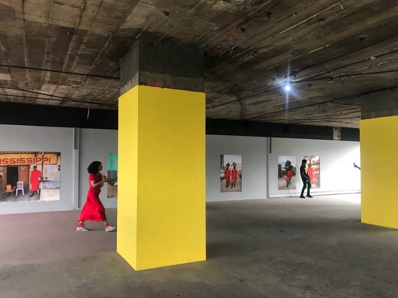 A woman dressed in bright red walks across an art exhibition. There is a large yellow column in the middle of the image. In the background of the image, there are photographs of women dressed in red standing in various homes, storefronts, and outdoor locations.