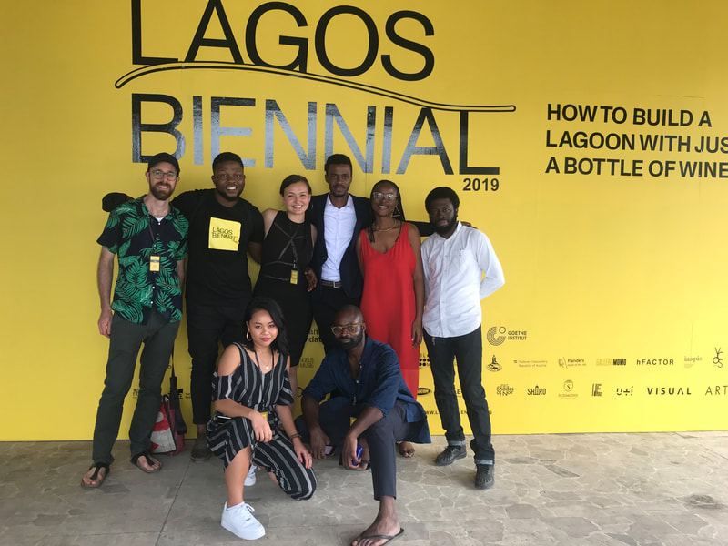 A group of eight people pose in front of a background with the words "Lagos Biennial 2019: How to Build a Lagoon with Just a Bottle of Wine?" Most people are dressed in black, with two people in white shirts, one person in a shirt patterned with leaves, and a person wearing a red dress.